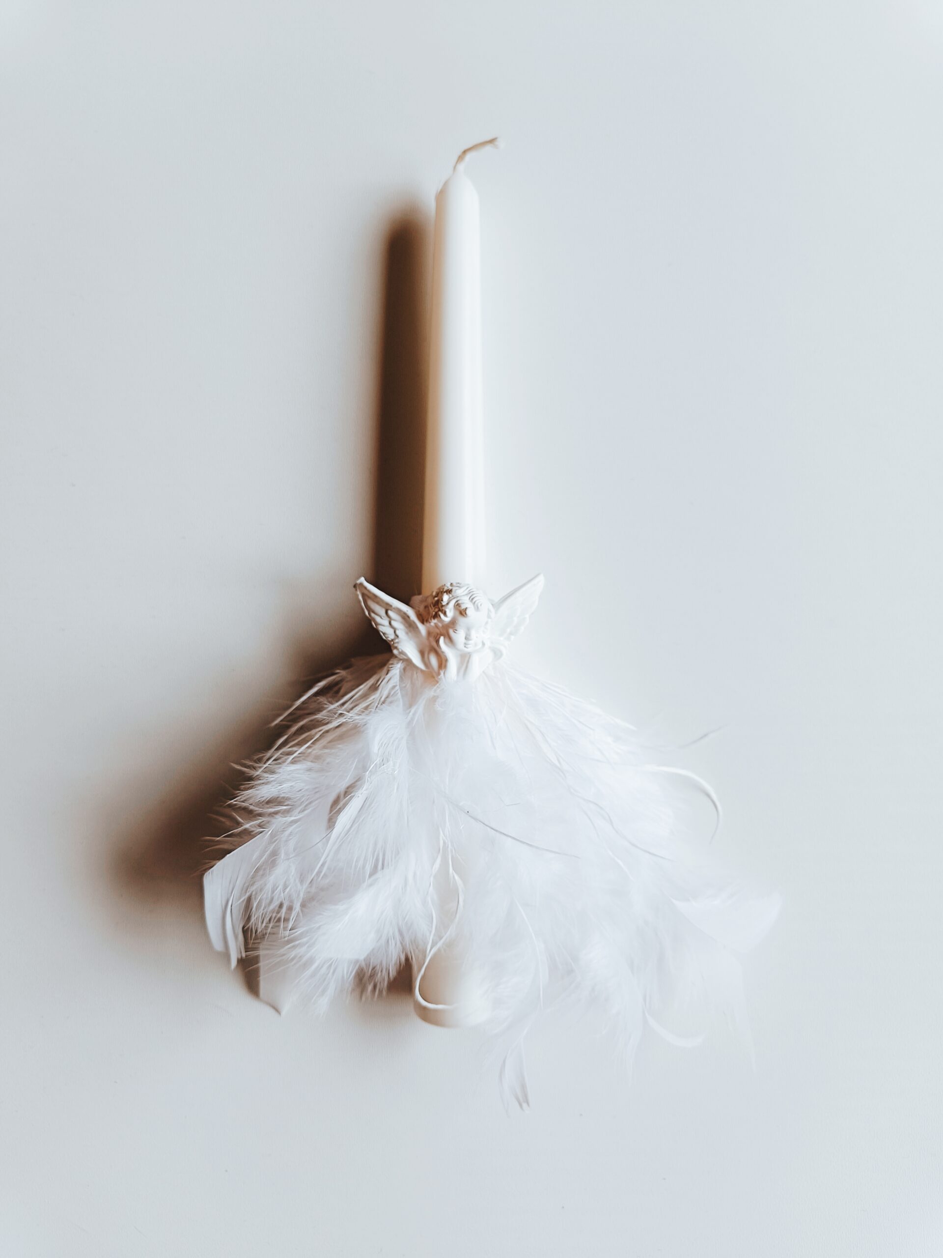 Žvakė dekoruota baltomis plunksnomis ir angeliuku